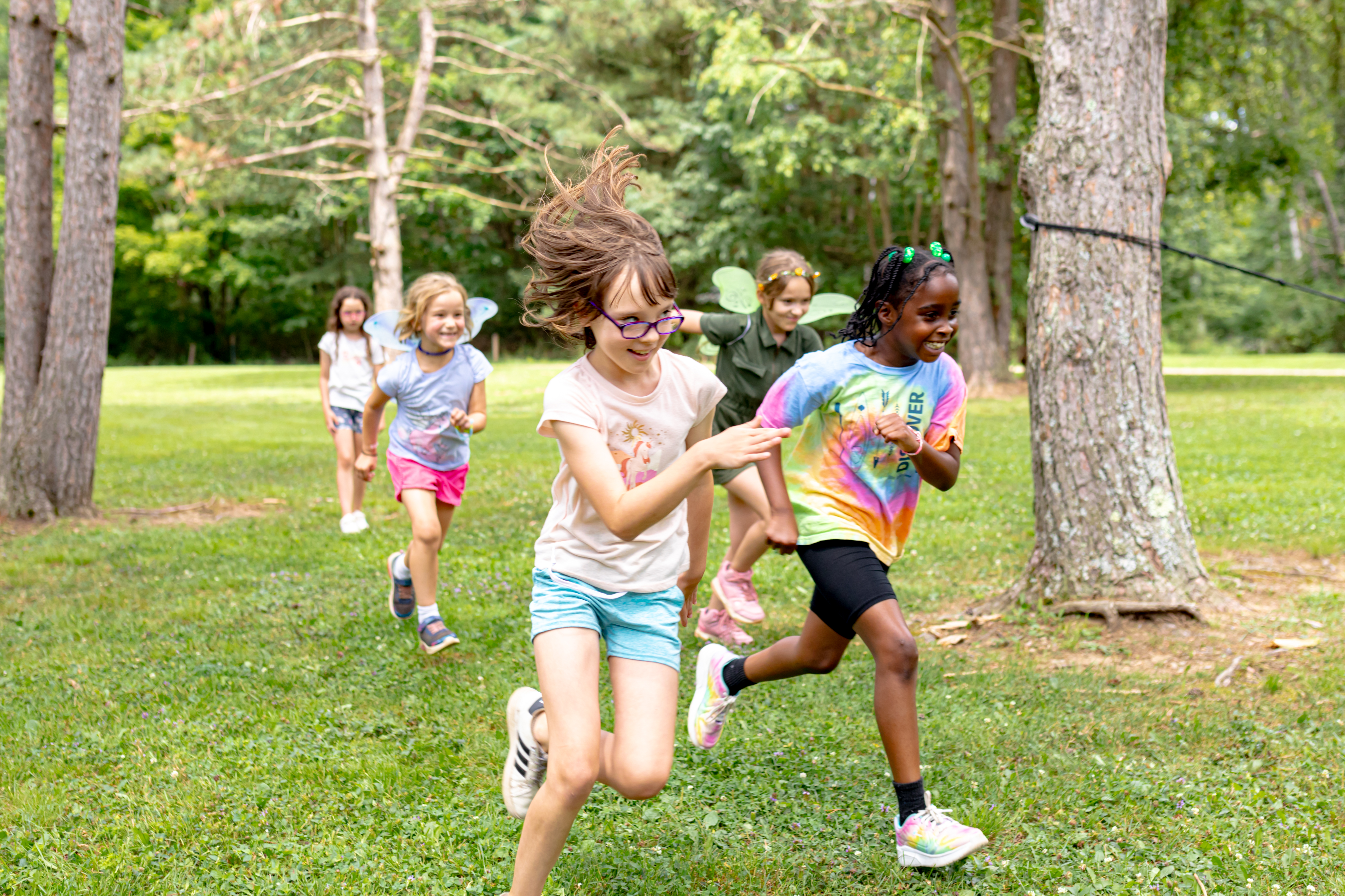  A group of Girl Scouts runs joyfully through a grassy area surrounded by trees, with two scouts in the front laughing as they race. 