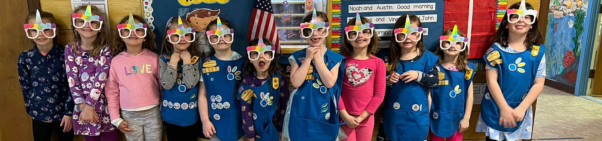  A group of 11 young girls standing in a classroom wearing glasses with rainbow unicorn horns on them 