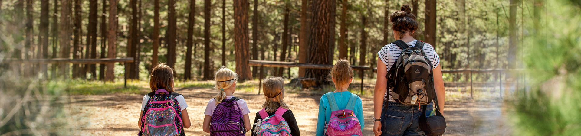  An adult volunteer and four young girls outdoors, each facing away from the camera and wearing a colorful backpack 