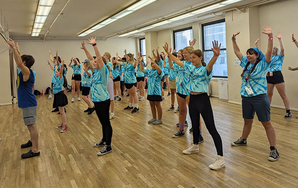 A group of teen girls all wearing blue tie dye shirts in a dance class with their arms up