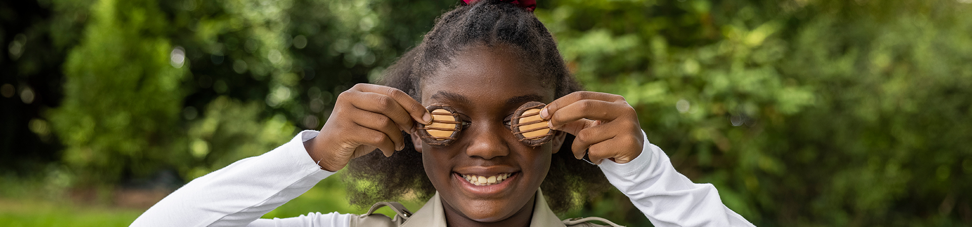  A young girl with a dark skin tone smiling for the camera and holding an Adventureful cookie over each eye 