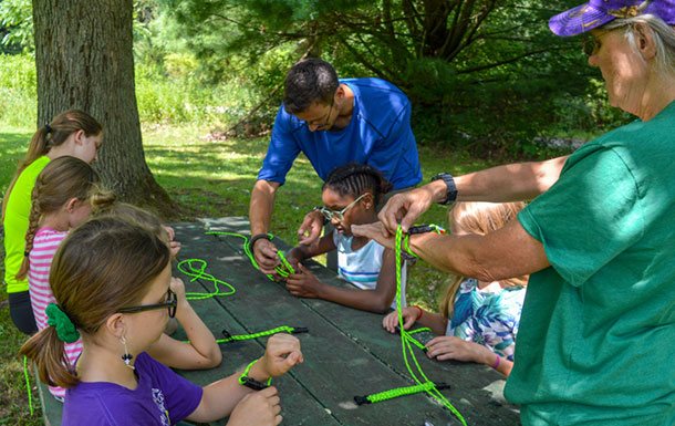 A group of girls sitting at a table outdoors weaving neon green ropes while adult volunteers help