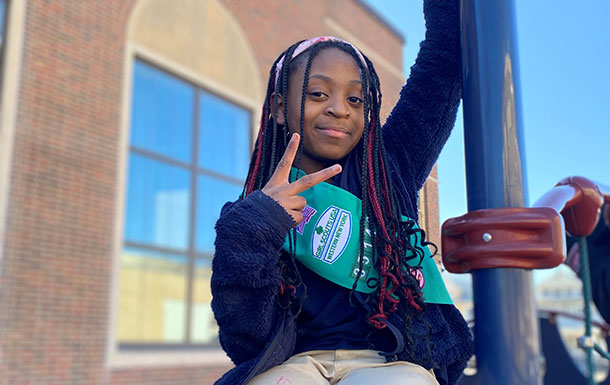 A smiling girl with a dark skin tone and long braids wearing her Girl Scout sash and holding up a peace sign