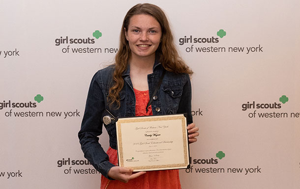 A smiling teen girl with a light skin tone and long brown hair wearing a black jacket and an orange dress and holding her Gold Award