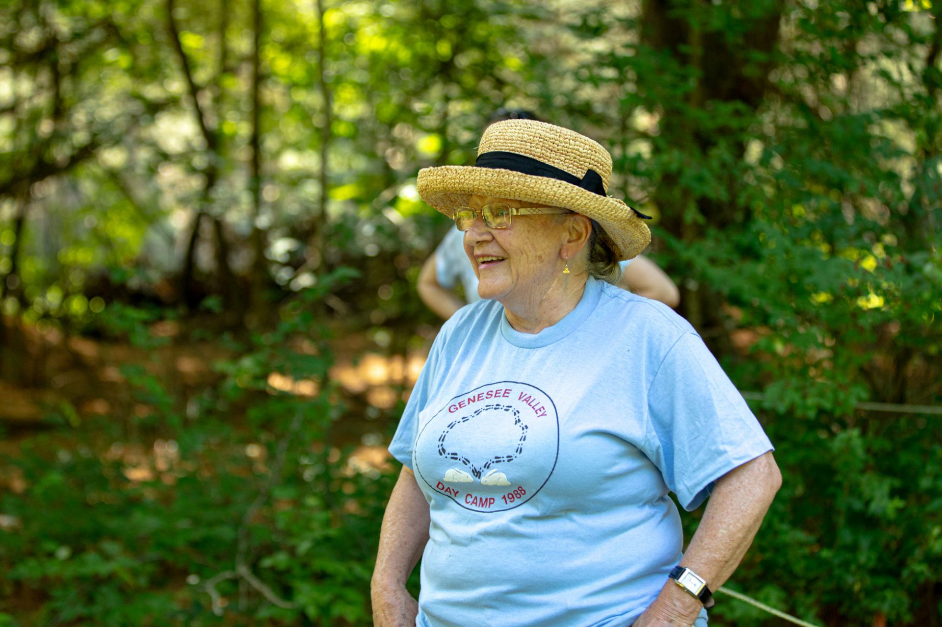 A smiling older woman with a light skin tone wearing a sunhat, glasses, and a blue T shirt that reads "Genesee Valley Day Camp"