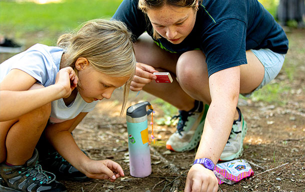 An adult woman with light skin tone and brown hair crouches on the ground to help a young girl with a light skin tone and blonde hair with her task