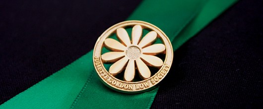 A gold circular pin with a daisy in the middle and the words "Juliette Gordon Low Society" inscribed on the border